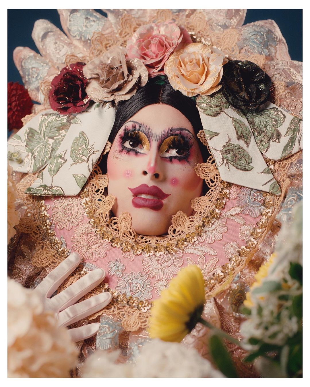 Portrait of a person with exaggerated makeup wearing an elaborate floral headpiece and lace ruff, posed with white-gloved hands and flowers.