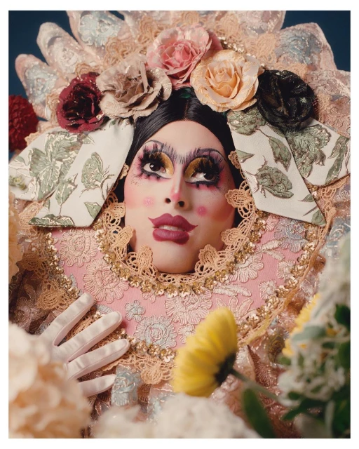 Portrait of a person with exaggerated makeup wearing an elaborate floral headpiece and lace ruff, posed with white-gloved hands and flowers.