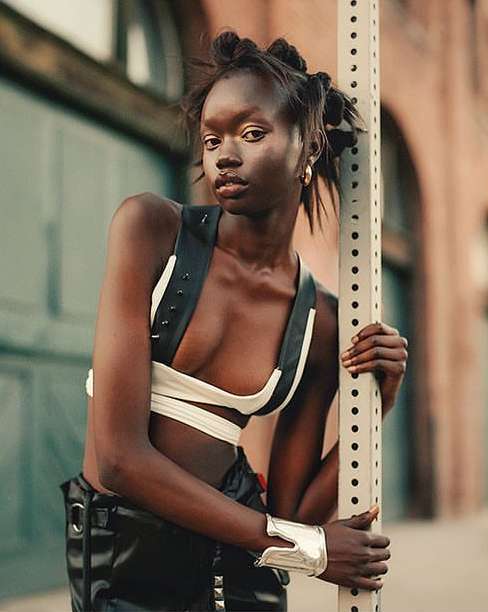 A striking portrait of a Black woman with stylishly knotted hair, posing in a black-and-white top and black pants, standing outdoors.