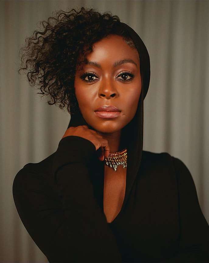 Headshot of a Black adult female wearing a black hooded dress and a gold necklace, gazing directly at the camera with a neutral expression.