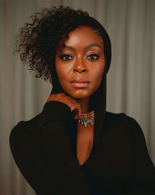 Headshot of a Black adult female wearing a black hooded dress and a gold necklace, gazing directly at the camera with a neutral expression.