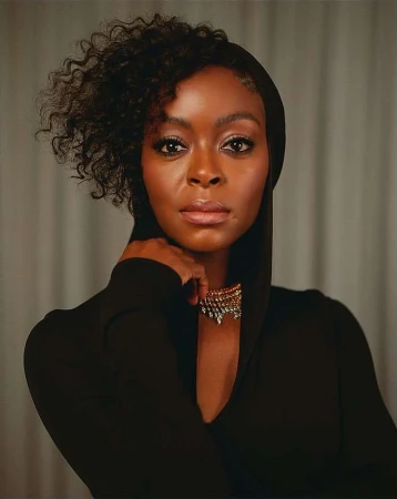 Headshot of a Black adult female wearing a black hooded dress and a gold necklace, gazing directly at the camera with a neutral expression.