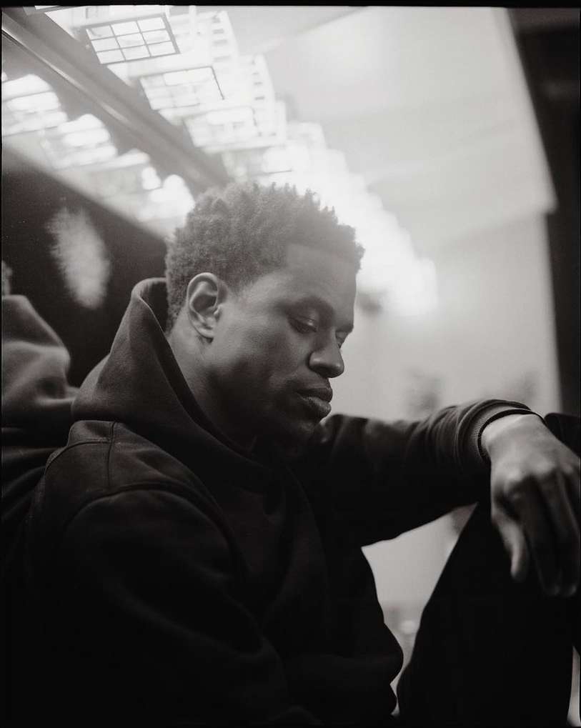 Black and white portrait of a thoughtful African American man in a hoodie, seated with his arms resting on his legs, indoor setting.