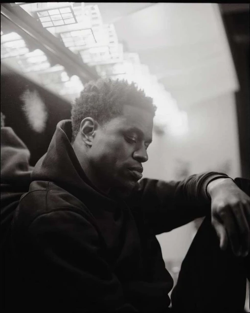 Black and white portrait of a thoughtful African American man in a hoodie, seated with his arms resting on his legs, indoor setting.