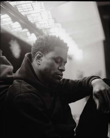 Black and white portrait of a thoughtful African American man in a hoodie, seated with his arms resting on his legs, indoor setting.