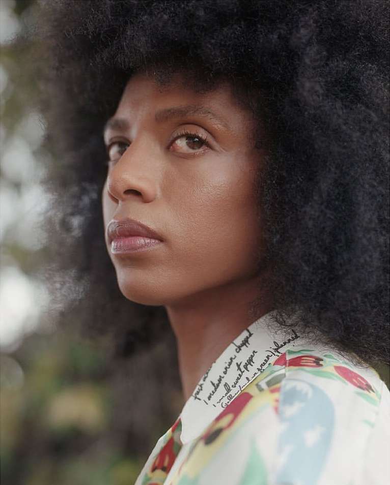 Close-up portrait of a woman with natural hair wearing a patterned shirt, set against a blurred background of greenery.