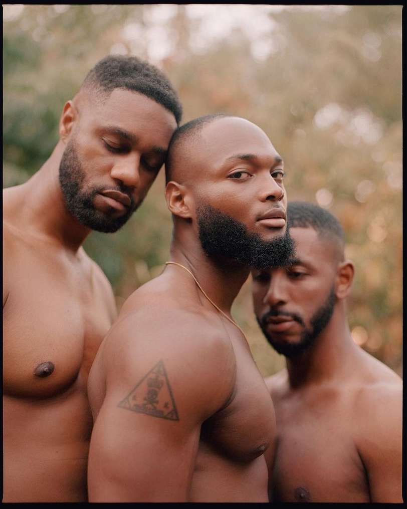 Portrait of three shirtless African-American men with beards in a natural outdoor setting, medium shot, warm tones, and shallow depth of field.