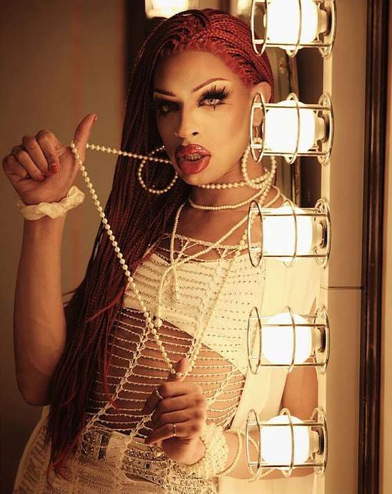 Person with auburn braided hair and dramatic makeup posing with jewelry against a backdrop of warm lighting and a light fixture.