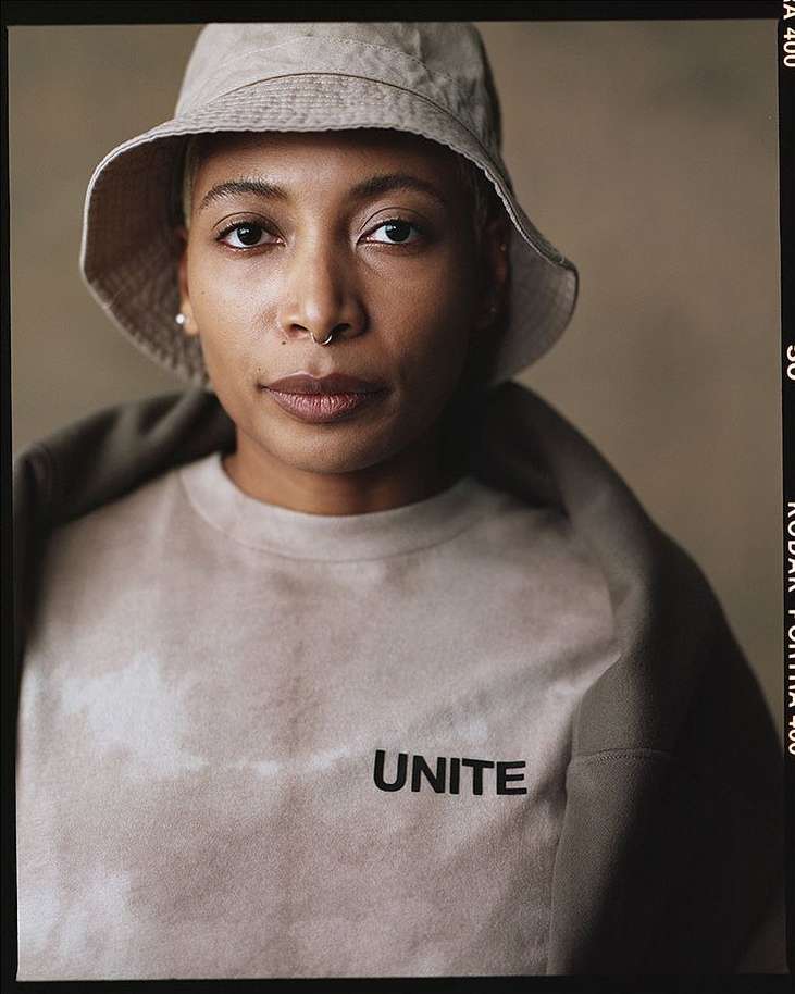 Close-up portrait of a young woman with short hair wearing a bucket hat and a tie-dye shirt with 'UNITE' printed on the front.