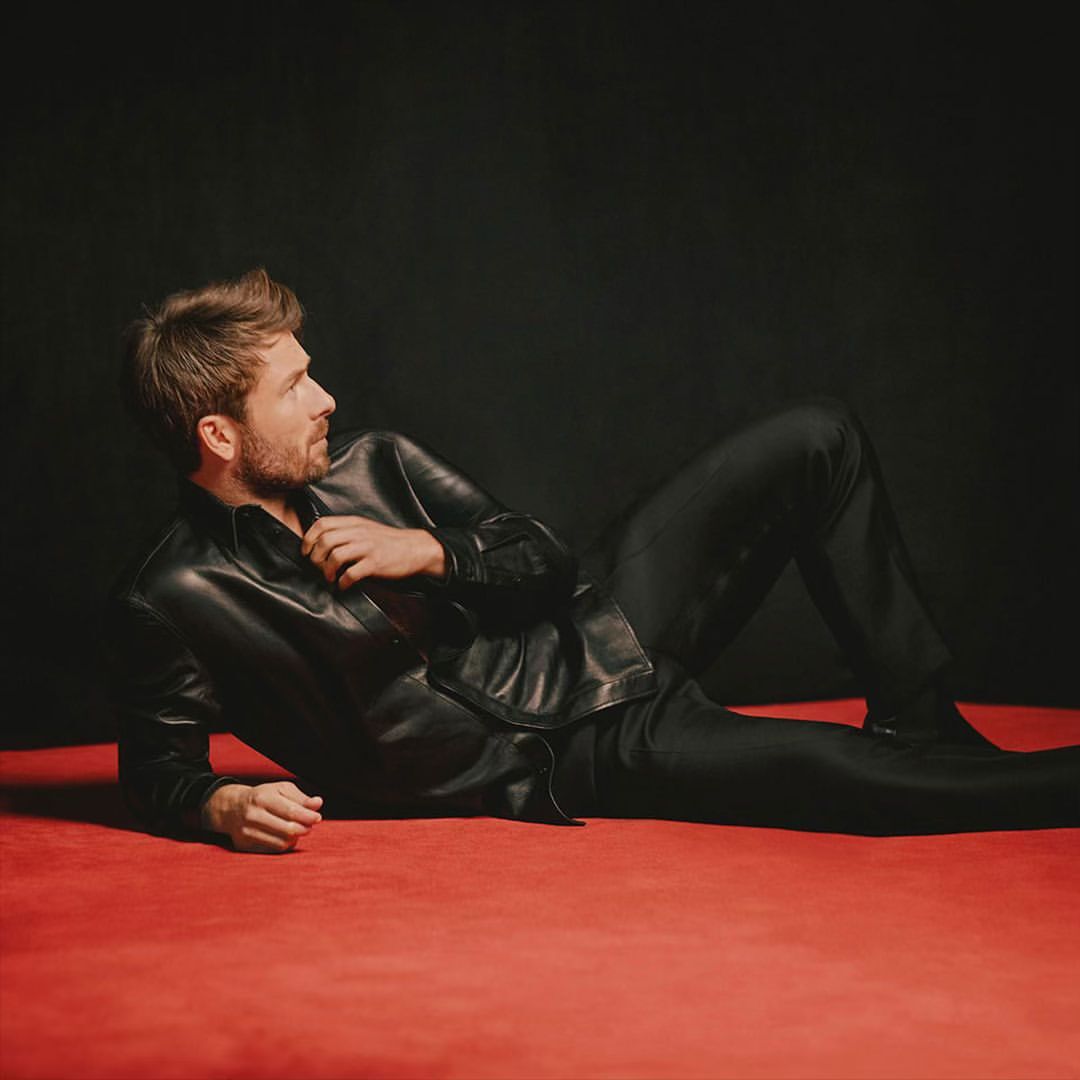 Man lying on a red surface wearing a black leather shirt and black pants, looking to the upper-left against a black background in studio lighting.