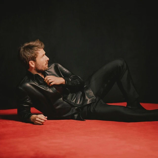 Man lying on a red surface wearing a black leather shirt and black pants, looking to the upper-left against a black background in studio lighting.