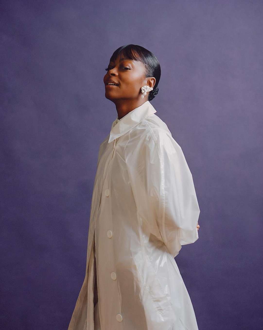 Portrait of a Black female model in a translucent white coat against a purple background, highlighted by a close-up studio shot.