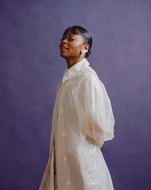 Portrait of a Black female model in a translucent white coat against a purple background, highlighted by a close-up studio shot.