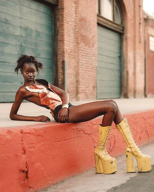 Fashion editorial shot of a Black female model in a sculptural top and platform boots, posing against an urban wall, capturing edgy style.