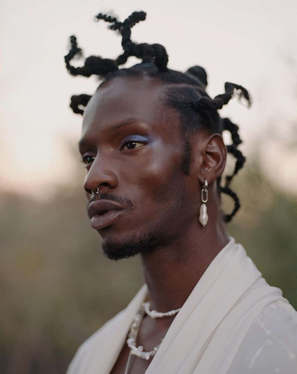 Close-up of an adult male with an elegant pearl necklace and statement earrings, styled hair, and neutral makeup against a soft outdoor background.