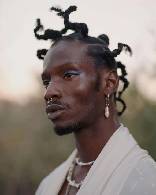 Close-up of an adult male with an elegant pearl necklace and statement earrings, styled hair, and neutral makeup against a soft outdoor background.