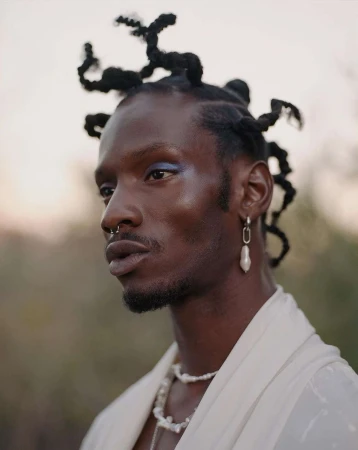 Close-up of an adult male with an elegant pearl necklace and statement earrings, styled hair, and neutral makeup against a soft outdoor background.