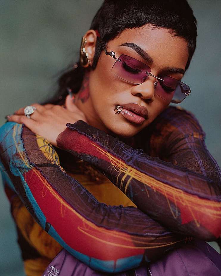 Close-up portrait of a woman wearing sunglasses, a patterned long-sleeve shirt, large gold earrings and rings against a green background.