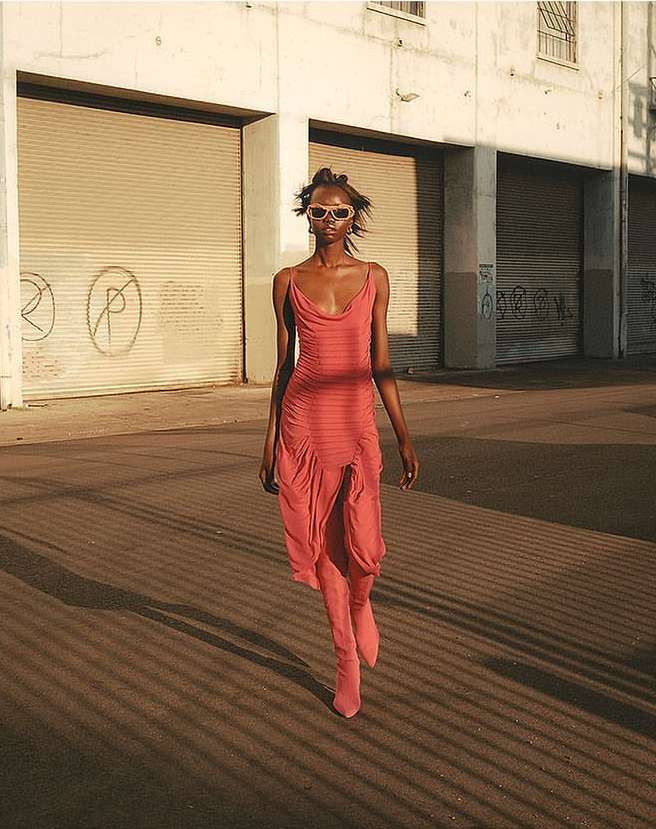 Fashion shot of a Black woman in a coral dress and matching boots walking on a street in front of a graffiti-covered building.