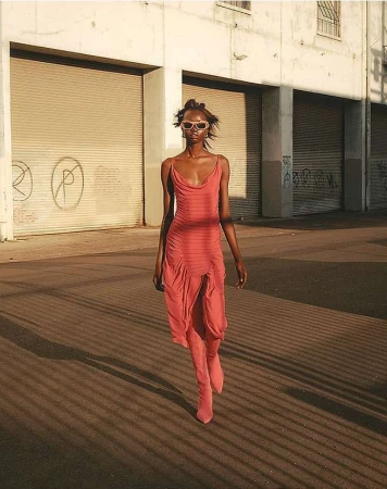 Fashion shot of a Black woman in a coral dress and matching boots walking on a street in front of a graffiti-covered building.