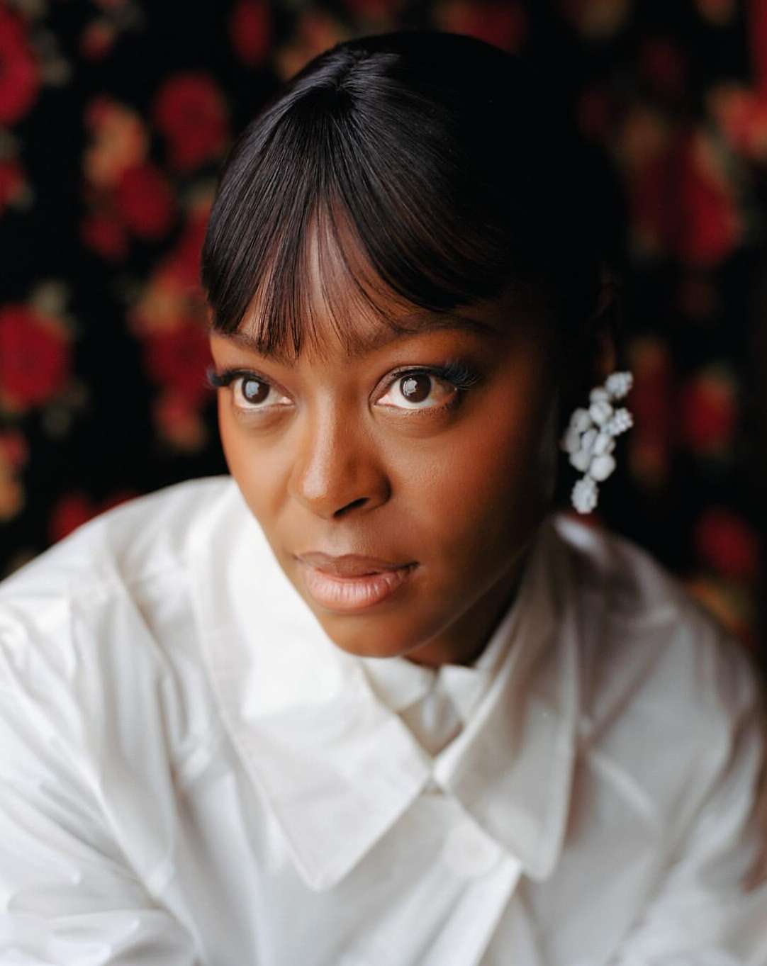 Close-up portrait of a Black woman wearing a white collared shirt and decorative floral earrings against a dark patterned background.