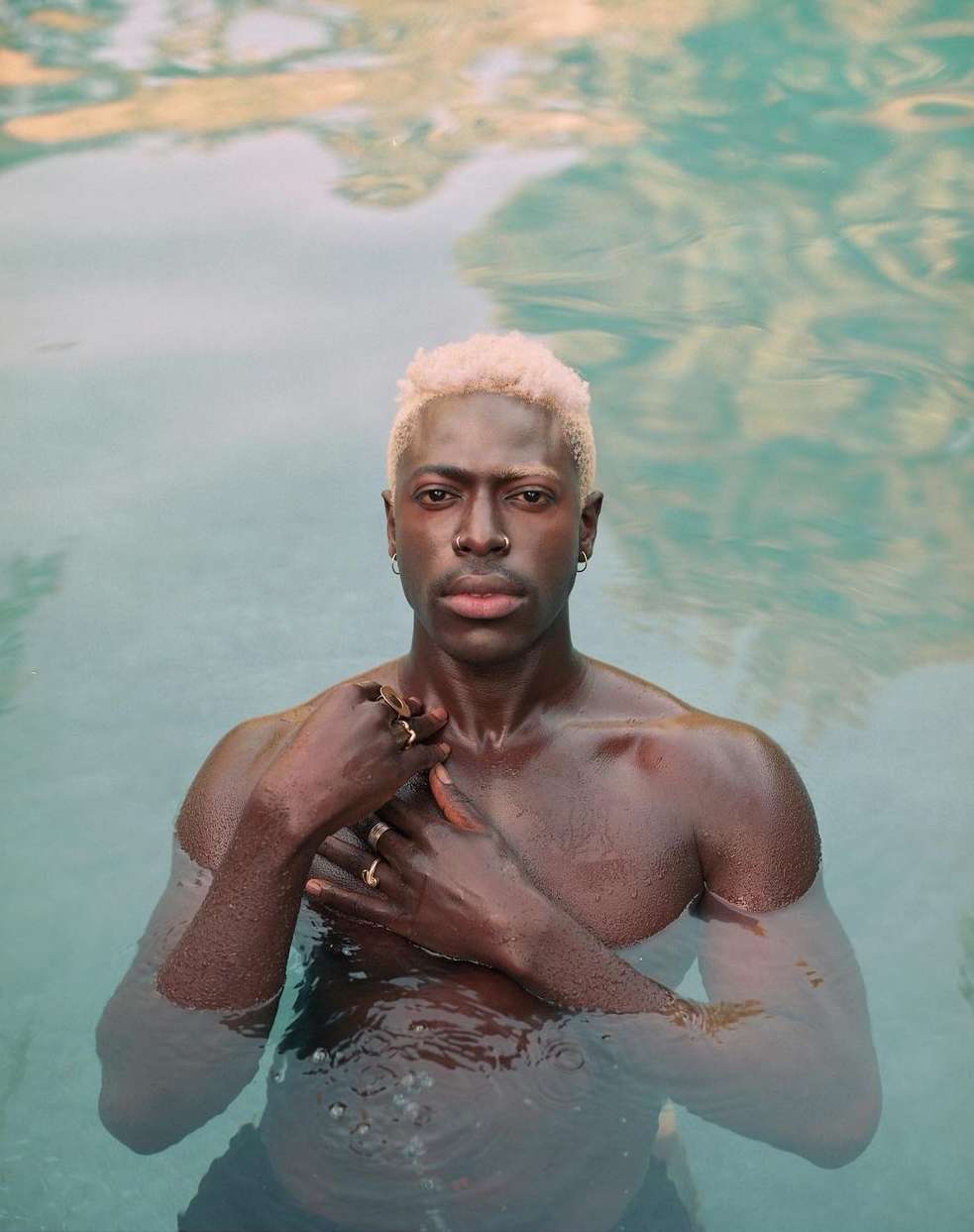 Adult male with blond hair stands in water. He wears rings and earrings. The background features reflections of sunlight and sky.