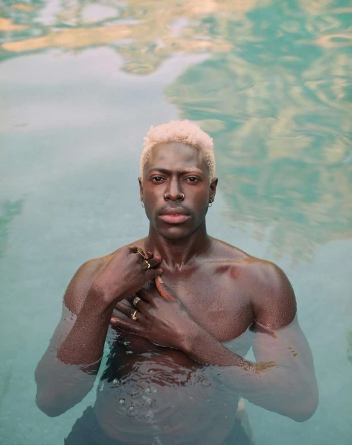 Adult male with blond hair stands in water. He wears rings and earrings. The background features reflections of sunlight and sky.