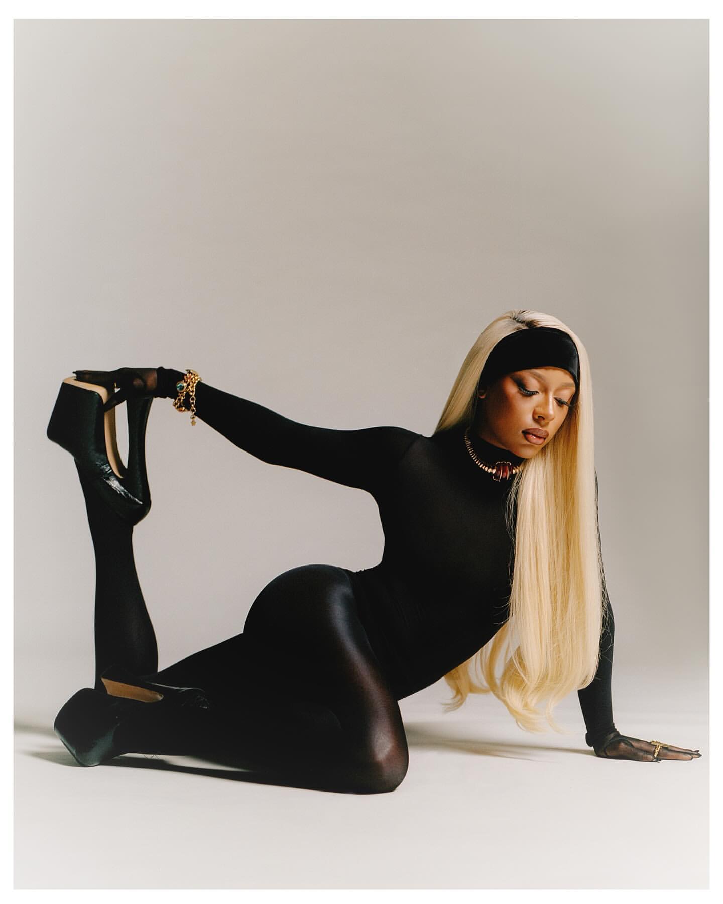 A woman in a black bodysuit and high heels is striking a pose on a white surface. The image is shot in a photo studio.