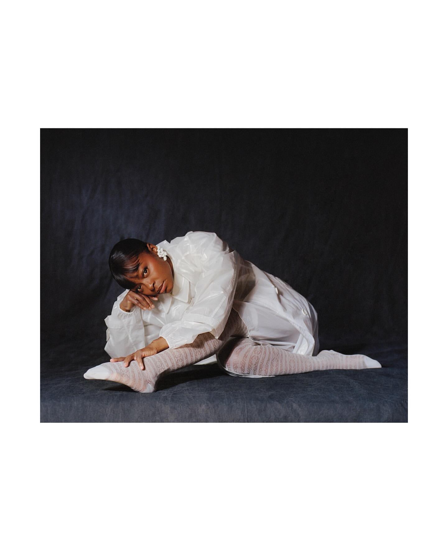 An adult woman poses in a glossy white coat and sheer hosiery in a studio with a dark grey background, resting on her elbow.