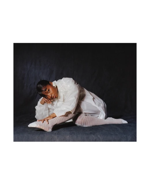 An adult woman poses in a glossy white coat and sheer hosiery in a studio with a dark grey background, resting on her elbow.