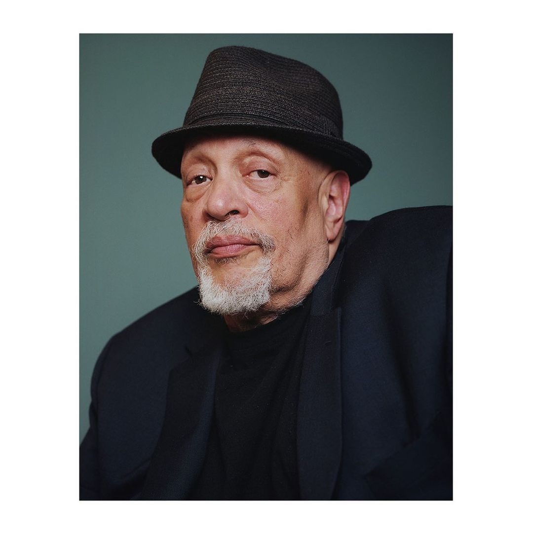 Close-up portrait of an older adult male with a grey beard, wearing a dark fedora hat and blazer against a muted green backdrop.