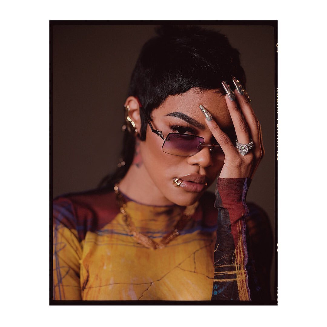 Stylish woman with short hair, sunglasses, and a patterned top poses indoors with manicured nails and gold jewelry.
