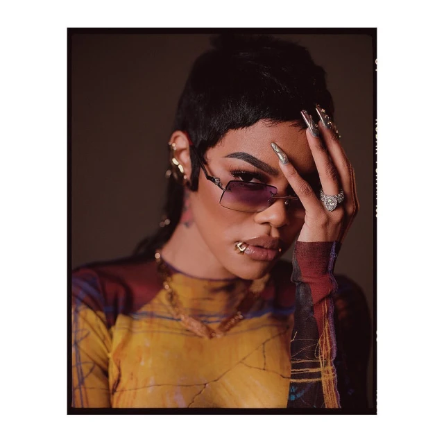 Stylish woman with short hair, sunglasses, and a patterned top poses indoors with manicured nails and gold jewelry.