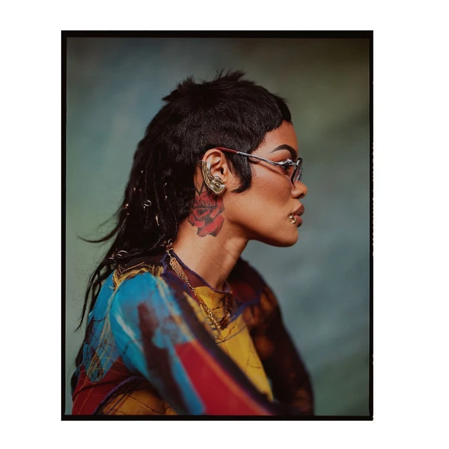 Stylish adult female poses in a side-profile shot, wearing sunglasses, jewelry, and a patterned top against a blurred background.