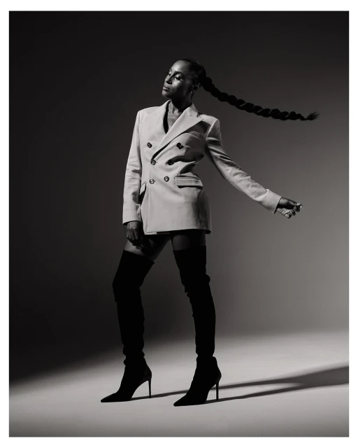 Black woman in an off-white blazer dress and black boots poses in a studio setting. Braided hair detail. Fashion editorial shot.