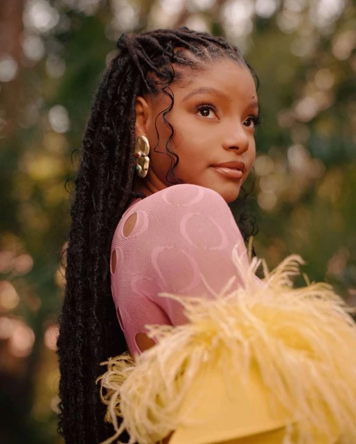 Portrait of an African American woman with long braids, wearing a pink mesh top and yellow feathered garment, in a natural outdoor setting.