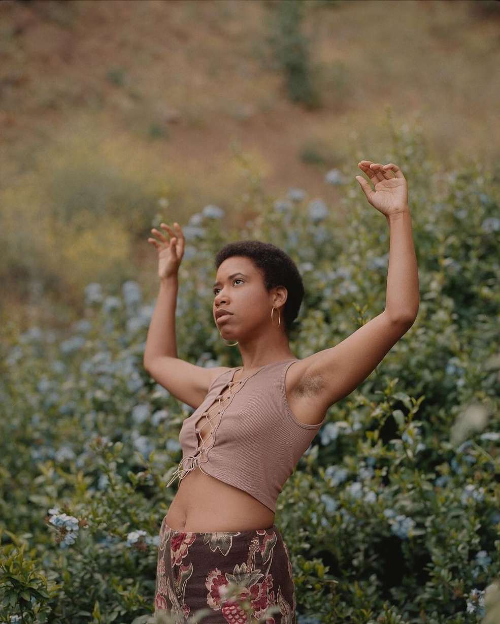 Adult woman stands among flowering bushes with her arms raised. The natural light reveals a stylish top and a printed skirt.
