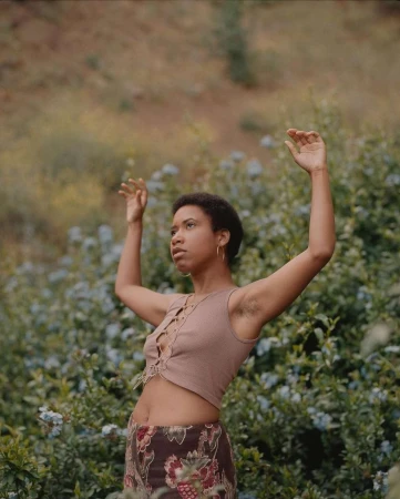 Adult woman stands among flowering bushes with her arms raised. The natural light reveals a stylish top and a printed skirt.