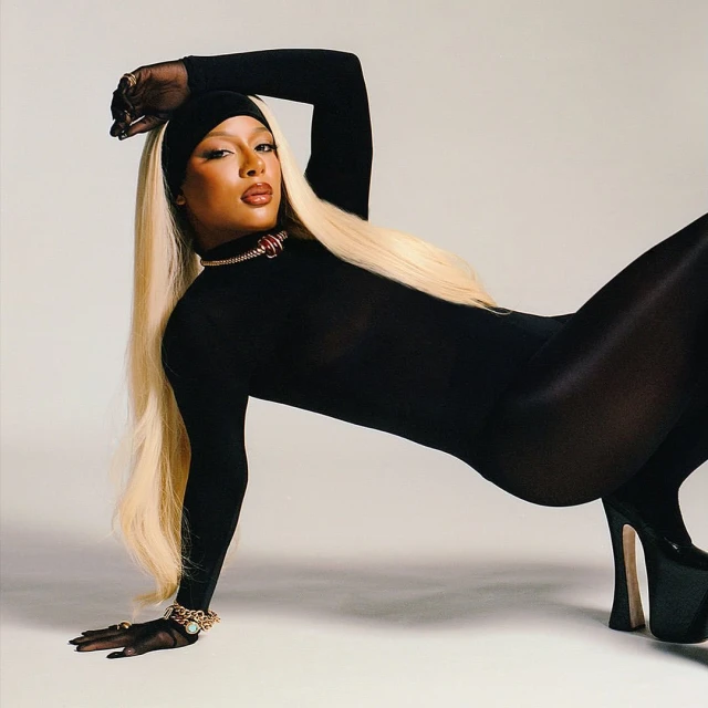 Adult female fashion model in a black bodysuit, headband, and platform heels posing indoors against a white background in a studio shot.