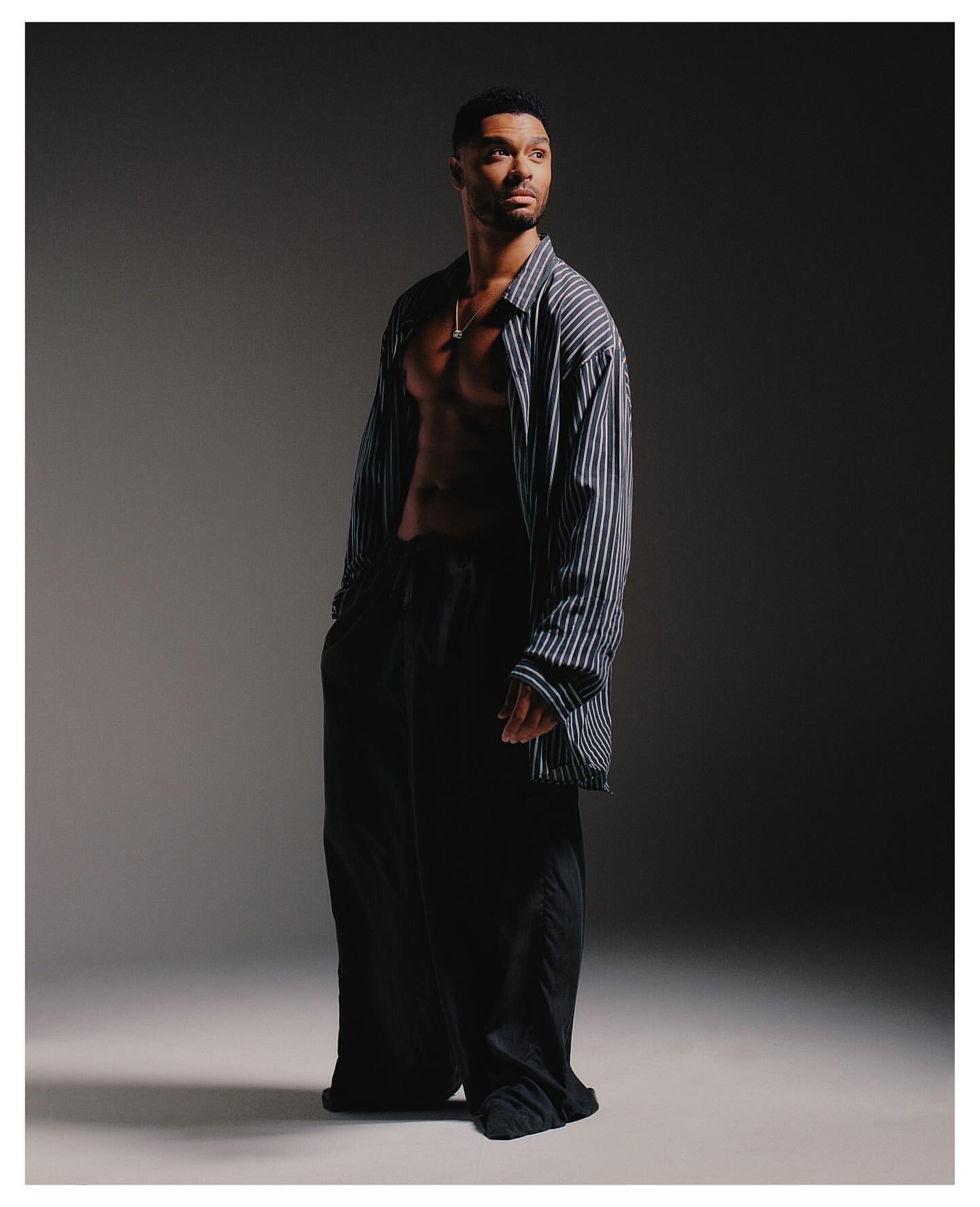 Portrait of a Black adult male model wearing a blue striped oversized shirt and black trousers in a photo studio with a gray backdrop.