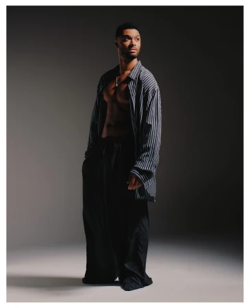 Portrait of a Black adult male model wearing a blue striped oversized shirt and black trousers in a photo studio with a gray backdrop.