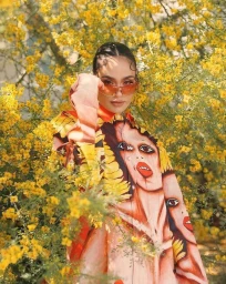 Woman wearing tinted sunglasses and a patterned shirt posing amidst bright yellow blooming foliage.