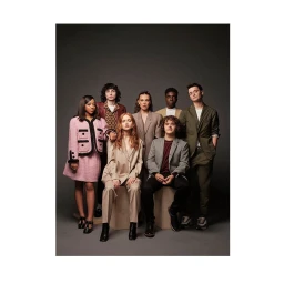 Group portrait of seven young actors standing and seated together against a dark grey studio background.