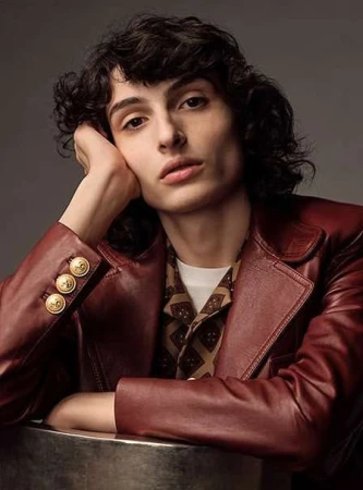 Young man with dark curly hair wearing a dark red leather blazer, resting his head on his hand against a grey studio backdrop.