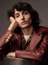 Young man with dark curly hair wearing a dark red leather blazer, resting his head on his hand against a grey studio backdrop.