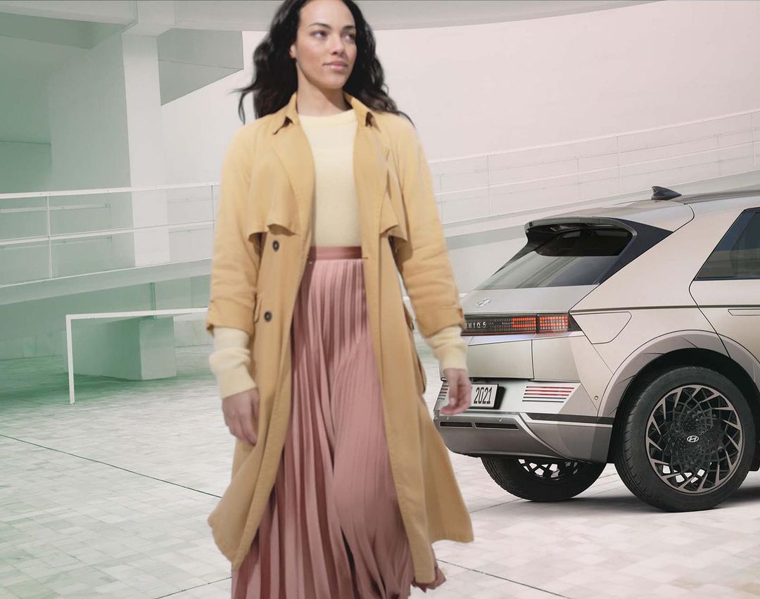 Adult female in trench coat walking near a silver 2023 Hyundai IONIQ 5 electric SUV in a modern, bright, indoor setting.