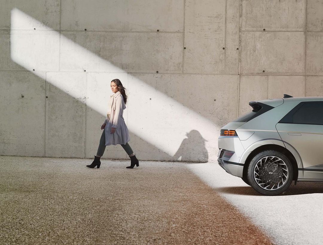 Woman in trench coat walking beside a silver Hyundai IONIQ 5 electric car in front of a concrete wall with sunlight.
