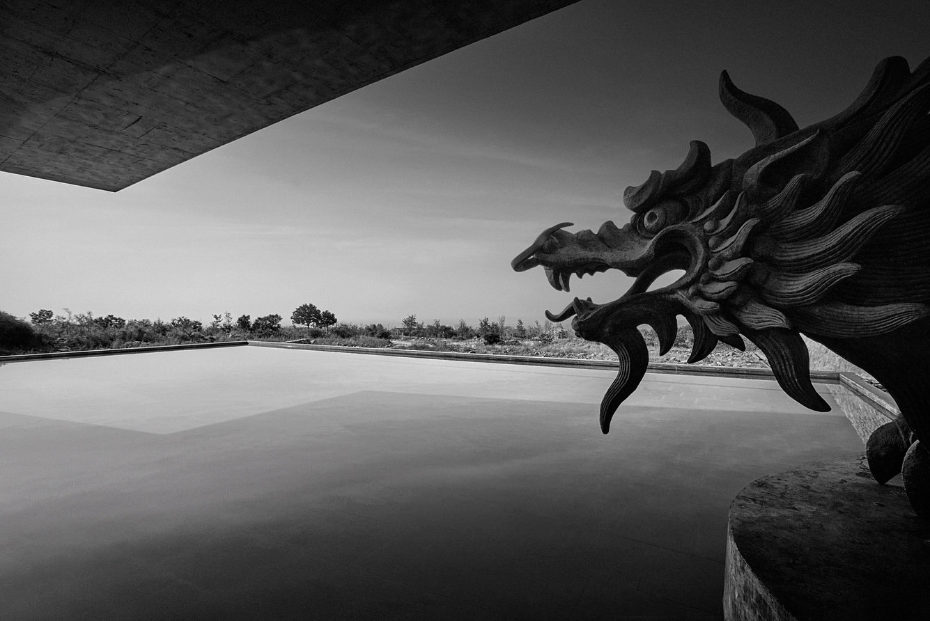 Monochrome photo of a dragon sculpture overlooking a reflective pool surrounded by greenery, captured beneath a concrete architectural overhang.