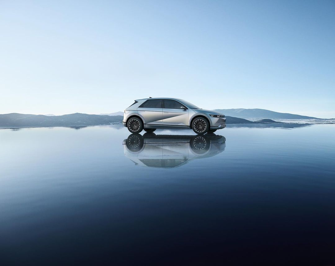 Silver Hyundai IONIQ 5 electric SUV reflected on a dark water surface against a light blue sky and distant mountains, showcasing its modern design.