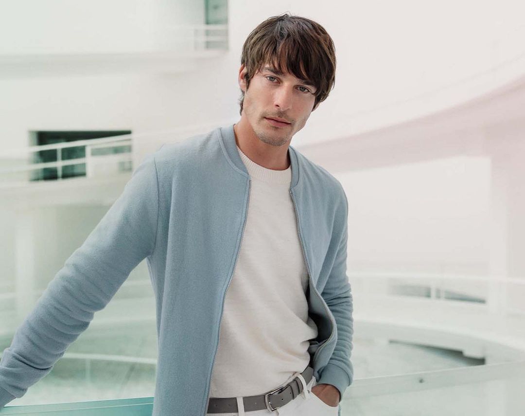 Adult male in a light blue bomber jacket and white jeans poses in a bright, airy interior with white walls and glass railings.